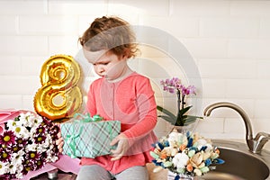 Cute funny little girl holding gift box sitting on kitchen table with bouquets of spring flowers and golden ballon 8 eight ,