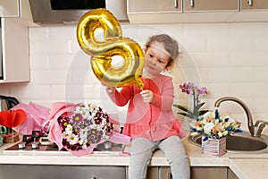 Cute funny little girl holding golden ballon 8 eight sitting on kitchen table with bouquets of spring flowers , greeting mother or