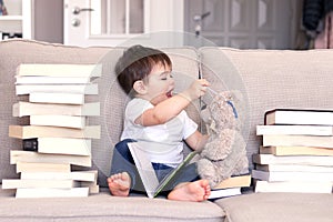 Cute funny playful little baby boy reading book and playing with teddy bear toy putting glasses on it sitting on sofa between pile