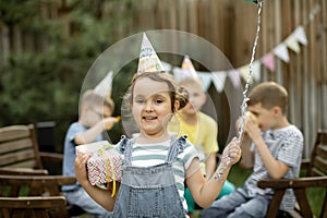 Cute funny six year old girl celebrating her birthday with family or friends in a backyard. Birthday party. Kid wearing party hat