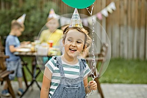 Cute funny six year old girl celebrating her birthday with family or friends in a backyard. Birthday party. Kid wearing party hat