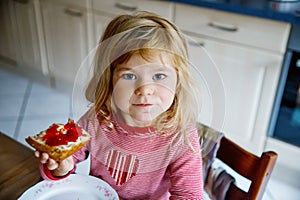 Cute funny toddler girl eats sweet bun for breakfast. Happy child eating bread roll with strawberry jam. Health food for
