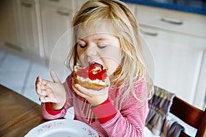 Cute funny toddler girl eats sweet bun for breakfast. Happy child eating bread roll with strawberry jam. Health food for