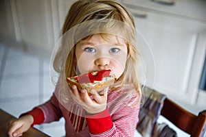 Cute funny toddler girl eats sweet bun for breakfast. Happy child eating bread roll with strawberry jam. Health food for