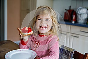 Cute funny toddler girl eats sweet bun for breakfast. Happy child eating bread roll with strawberry jam. Health food for