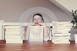 Cute funny tween girl in glasses sitting a table with pile of books looking sceptically and daydreaming of holidays.
