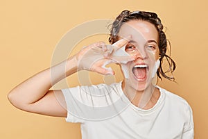 Cute funny woman wearing white T-shirt with hydrogel patches under eyes and shampoo foam on her head isolated over beige