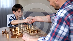 Cute genius kid winning chess game, handshaking with his proud father, hobby