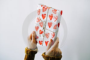 A cute gift in a woman`s hand wrapped in homemade wrapping paper with red hearts tied with jute thread for Valentine`s day on a