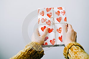 A cute gift in a woman`s hand wrapped in homemade wrapping paper with red hearts tied with jute thread for Valentine`s day on a