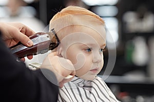 Cute ginger baby boy with blue eyes in a barber shop having haircut by hairdresser. Hands of stylist with tools.