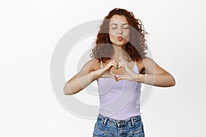 Cute ginger girl with curly hairstyle, kissing, pucker lips and showing heart, I love you gesture, standing over white