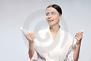 Cute girl in bathrobe holding bottle of cosmetic cream and pore cleansing brush