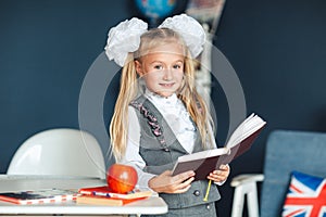 Cute girl blondy in school uniform with white bows standing near the desk and studying in the classroom. Education and school