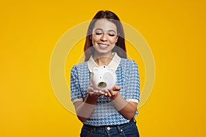 Cute girl in casual blue shirt holding white piggy bank with lots of money against yellow background