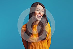 Cute girl in casual orange jumper smiling broadly with closed eyes against blue