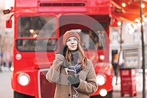 Cute girl coat and hats stand on the background of the red bus,listen music in the headphones
