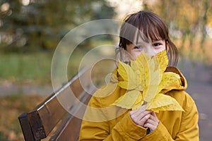 Cute girl in coat sitting on the bench. The girl holds an armful of leaves