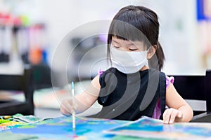 Cute girl is coloring on canvas. Kid wear white mask and black aprons uniform while sitting on wooden chair to make craft.