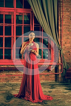 Cute girl with a Cup and a plate standing at the red big window in the evening