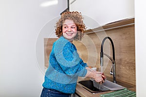 Cute girl with curly red hair washing apple in kitchen sink. Young Woman cleaning her fruit with water under the tap. Daily intake