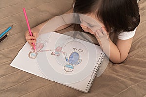 Cute girl draws a pencil drawing. A child learns to draw people. The child lies on the bed and draws with a pencil