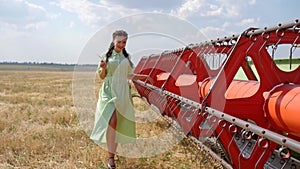 Cute girl in dress going in slow motion near agricultural machinery on background golden field