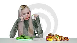 A cute girl eats greens and looks sadly at fast food on a white background. Diet. The concept of healthy and unhealthy