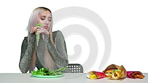 A cute girl eats greens and looks sadly at fast food on a white background. Diet. The concept of healthy and unhealthy