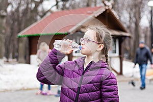 A cute girl in glasses drinks water from a bottle bought in a food truck in a city park. Takeaway food