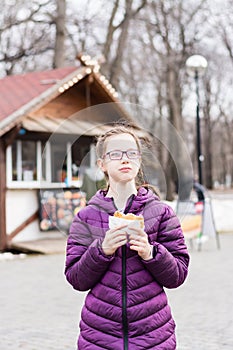 Cute girl with glasses eats a pie bought in a food truck in a city park. Takeaway food