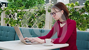 Cute girl in glasses is typing on laptop in cafe