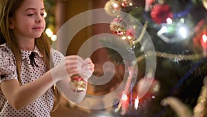 Cute girl hangs a decoration ball at the Christmas tree sitting on the floor in the room