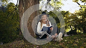 Cute girl in the hat reading book while sitting on the grass in the park.