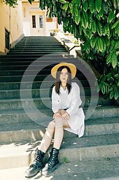 a cute girl in a hat is sitting on the stairs of the old town. Brunette in a white dress walks the street