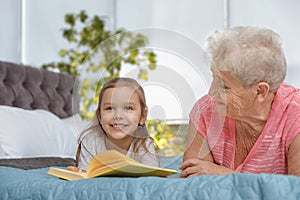 Cute girl and her grandmother reading book on bed