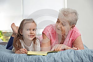Cute girl and her grandmother reading book on bed