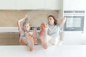 Cute girl and her mother are smiling while eating ice cream in the kitchen with legs on a table