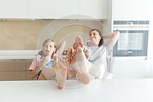 Cute girl and her mother are smiling while eating ice cream in the kitchen with legs on a table