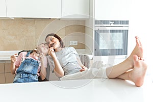 Cute girl and her mother are smiling while eating ice cream in the kitchen with legs on a table