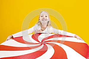 A cute girl holding a huge candy in her hands on a yellow background. Sweet gift. Big lollipop. Christmas sweetness