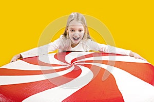A cute girl is holding a huge red and white candy on a yellow background. Sweet gift. Large lollipop. Christmas sweetness