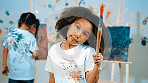 Cute girl holding painted brush while student drawing canvas behind. Erudition.