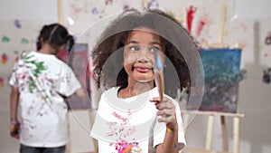 Cute girl holding painted brush while student drawing canvas behind. Erudition.