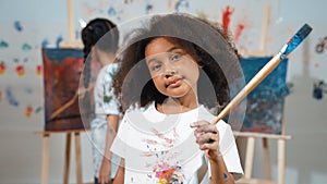Cute girl holding painted brush while student drawing canvas behind. Erudition.