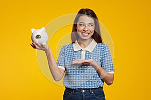 Cute girl holding and pointing at white piggy bank with lots of money isolated over yellow backdrop