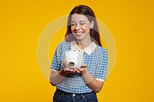 Cute girl holding white piggy bank while smiling broadly with closed eyes against yellow background