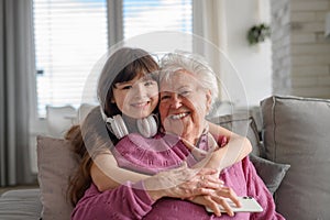 Cute girl hugging gradmother from behind, looking at camera, smiling. Portrait of an elderly woman spending time with