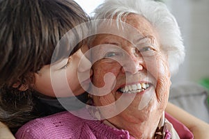 Cute girl hugging and kissing gradmother on cheek from behind. Portrait of an elderly woman spending time with