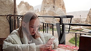 Cute Girl In Jacket Sits at Table in Open Terrace Cafe Restaurant and Type Text on Smart Phone Mobile.